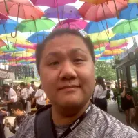 front-facing mugshot of Brandon Cha staring at you the user with colourful, opened, umbrellas hanging in the sky at the Stackt (sic) market in Toronto, Canada